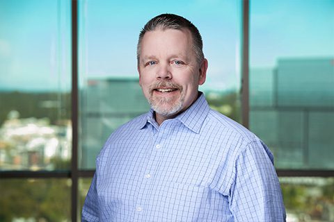 Man in a blue checked shirt smiles in front of a bright window with a blurred city view.