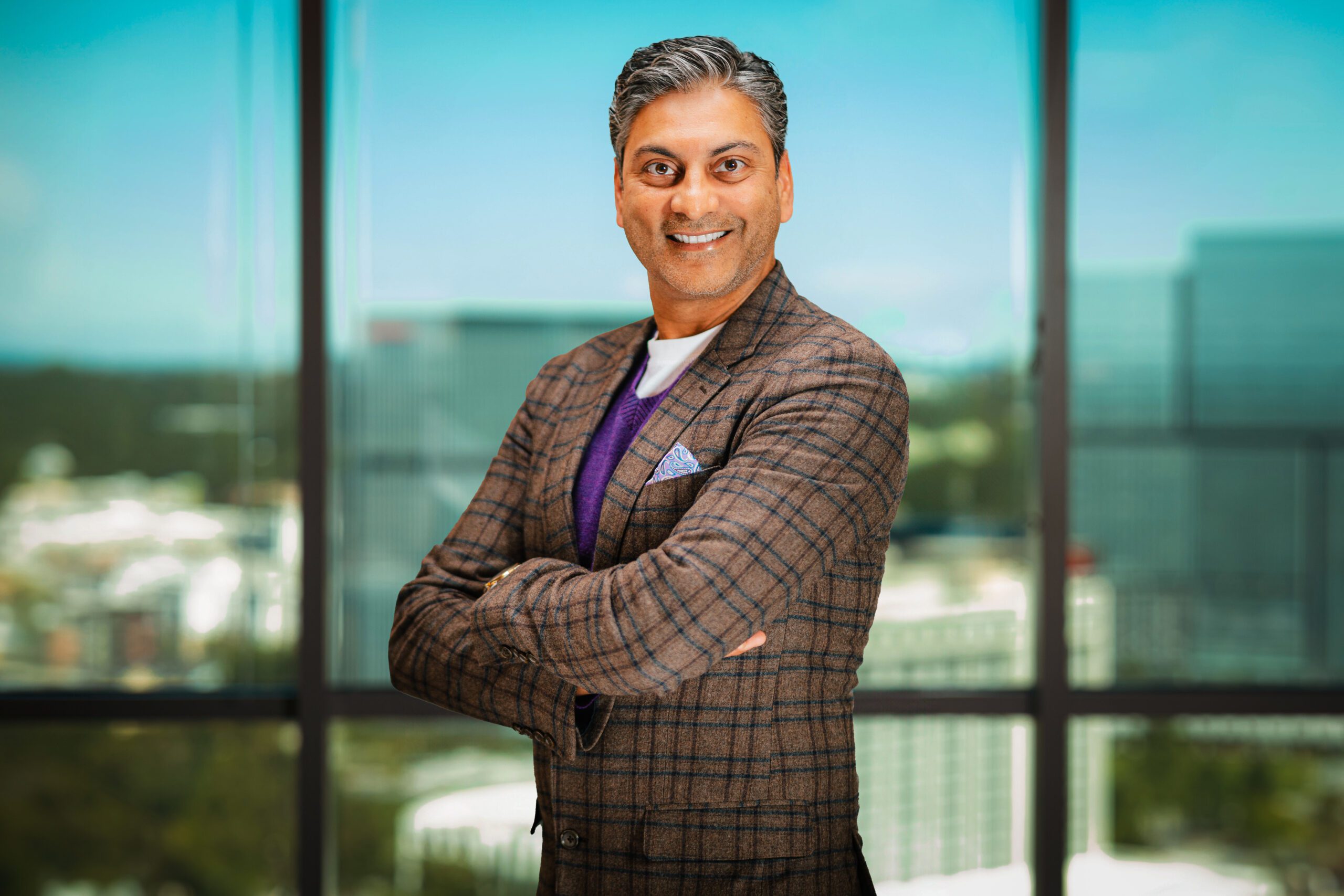 A man in a plaid blazer stands with arms crossed, smiling, in front of large windows overlooking a cityscape.