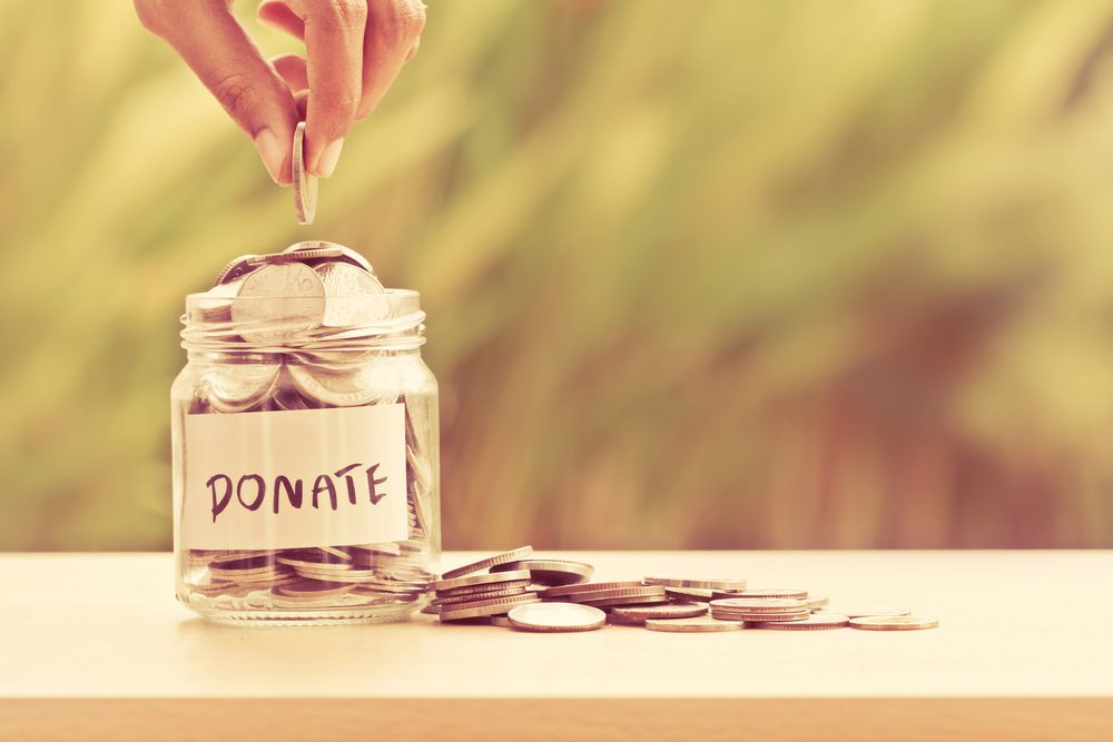A hand drops a coin into a glass jar labeled DONATE, with coins inside the jar and scattered on the surface beside it.