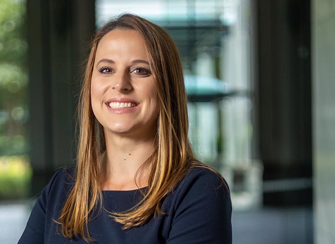 A woman with long brown hair wearing a navy blue top smiles at the camera in an indoor setting with blurred background.