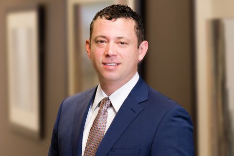 A man in a blue suit and patterned tie stands indoors near a blurred background with framed pictures on the wall.