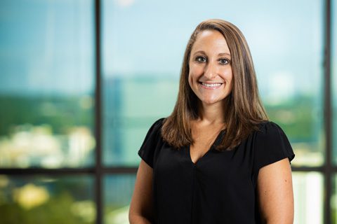 Woman with straight brown hair, wearing a black short-sleeve blouse, stands smiling in front of a large window with an urban landscape in the background.