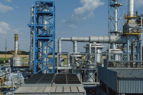 Industrial facility with large metal pipes, towers, and structures under a partly cloudy sky; fields visible in the background.