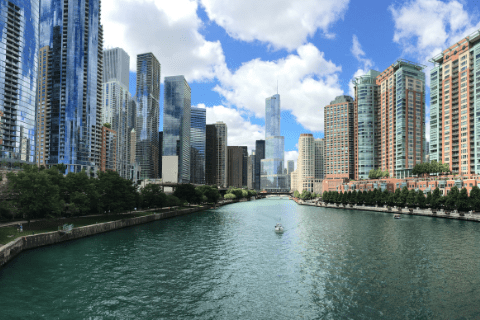 A cityscape featuring tall modern buildings on both sides of a river under a partly cloudy sky, with a small boat visible on the water.