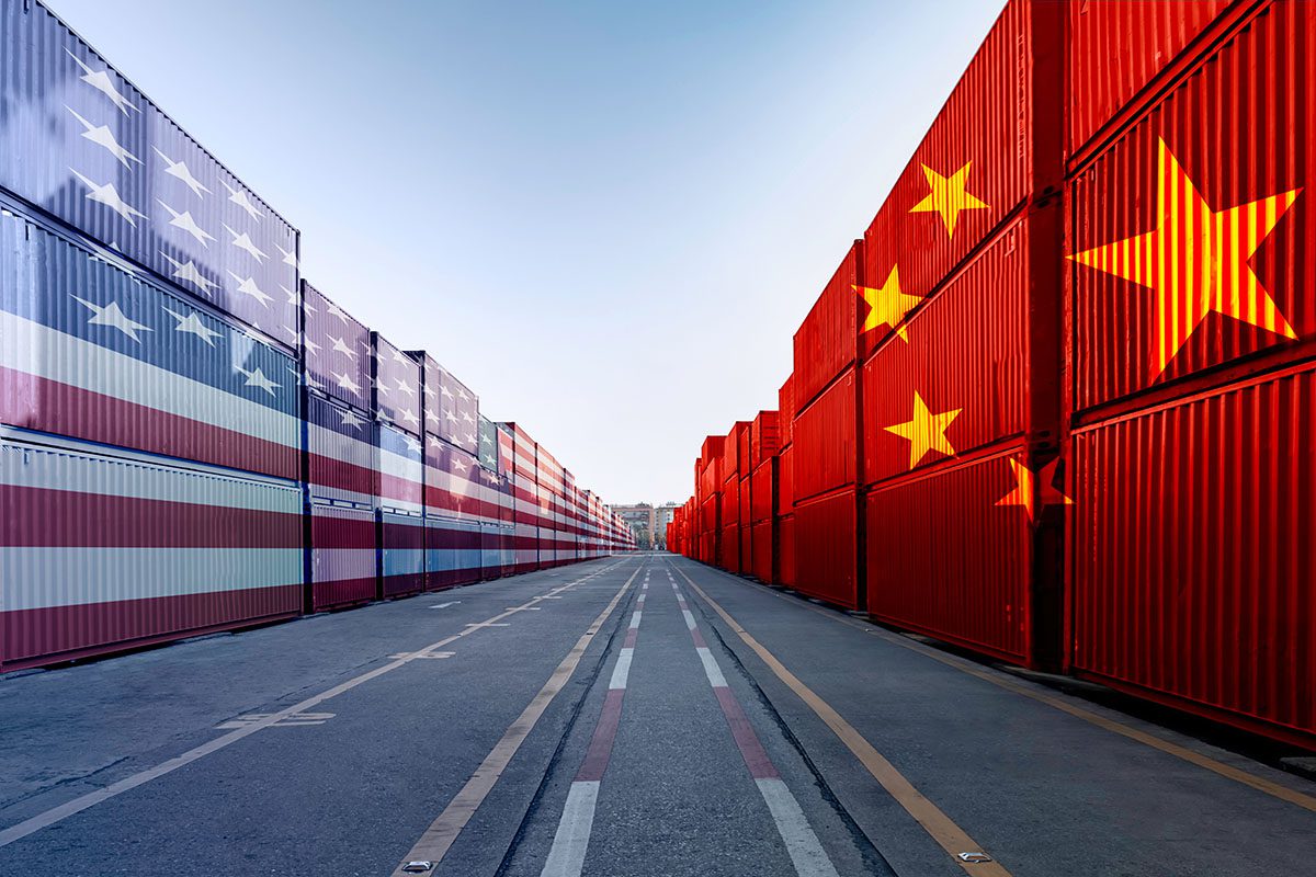 A row of shipping containers painted with the U.S. flag on the left and Chinese flag on the right lines an empty road under a clear sky.