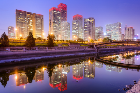 Tall modern skyscrapers with illuminated windows and pink lights on top are reflected in a calm river at dusk, with a bridge crossing the water and trees lining the riverside.