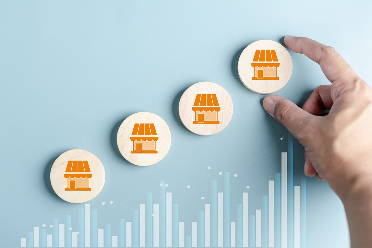 A hand arranges four wooden discs with store icons in ascending order, above a bar graph on a blue background, symbolizing business growth.
