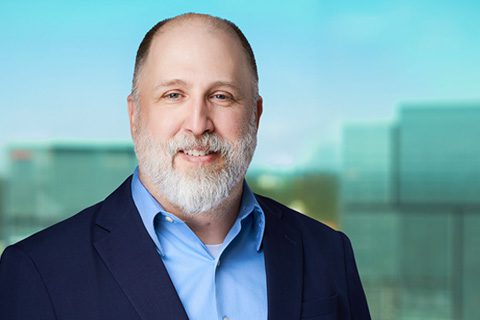 A middle-aged man with a beard, wearing a blue shirt and dark blazer, smiles in front of a blurred office background.