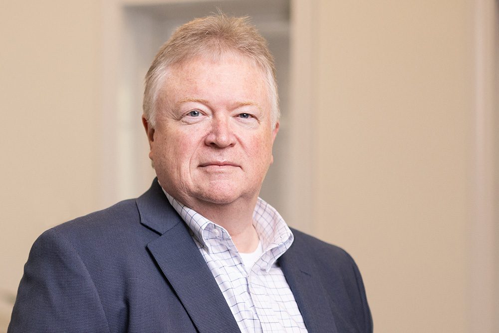 A middle-aged man with light hair wears a dark suit jacket and a white checkered shirt, standing indoors against a neutral background.