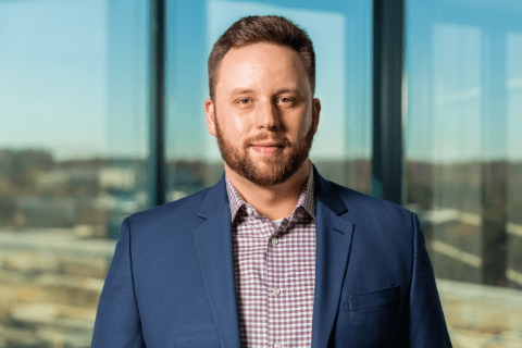 Man with short brown hair and beard wearing a blue suit jacket and checked shirt stands indoors in front of large windows with an urban view.