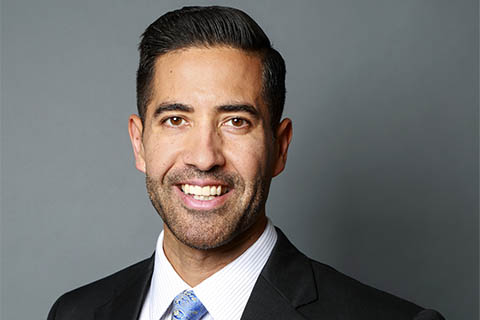 Man in a suit and tie smiling in front of a plain gray background.