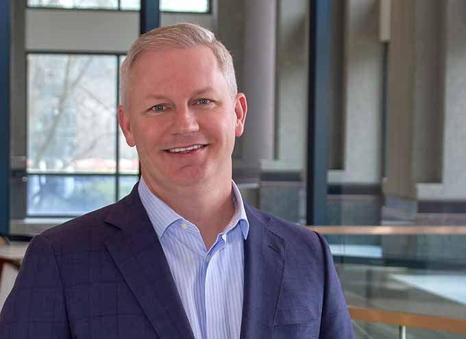 A man in a blue suit and striped shirt smiles while standing indoors with large windows in the background.