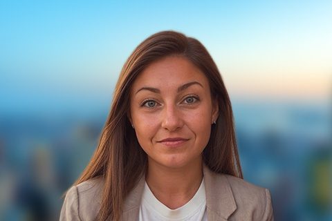 Woman with long brown hair and a blazer smiles in front of a bright window with a blurred city view