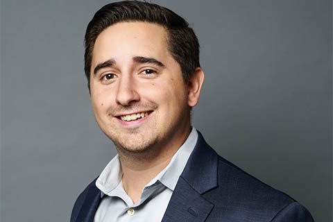 A man with short dark hair, wearing a navy blazer and light-colored shirt, smiles at the camera against a plain gray background.