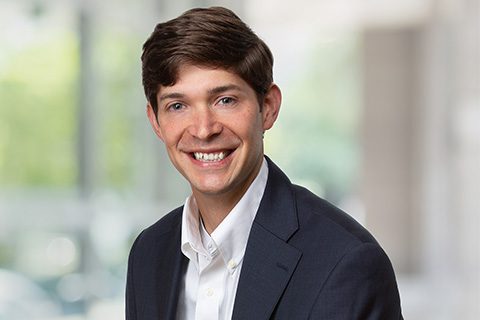 A man with short brown hair wearing a dark suit jacket and white shirt smiles at the camera against a blurred indoor background.