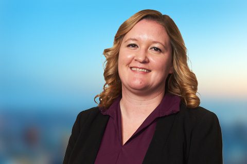 A woman with shoulder-length wavy blonde hair, wearing a dark blazer and maroon blouse, smiles in front of a blurred blue and orange background.