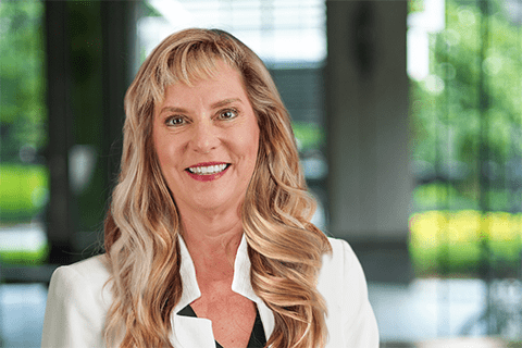 A woman with long blonde hair wearing a white blazer smiles at the camera in a bright, modern indoor setting with greenery visible outside.