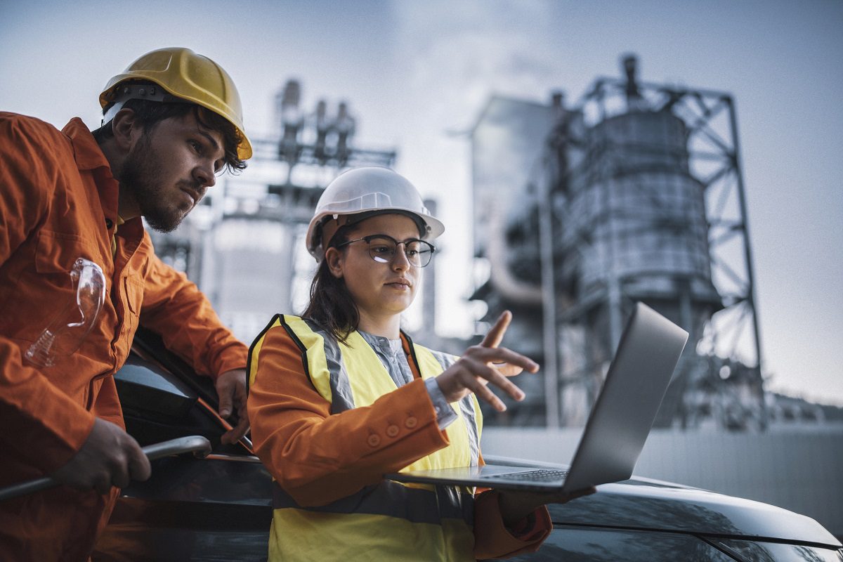 Two workers wearing safety gear review information on a laptop outdoors at an industrial facility with large metal structures in the background.