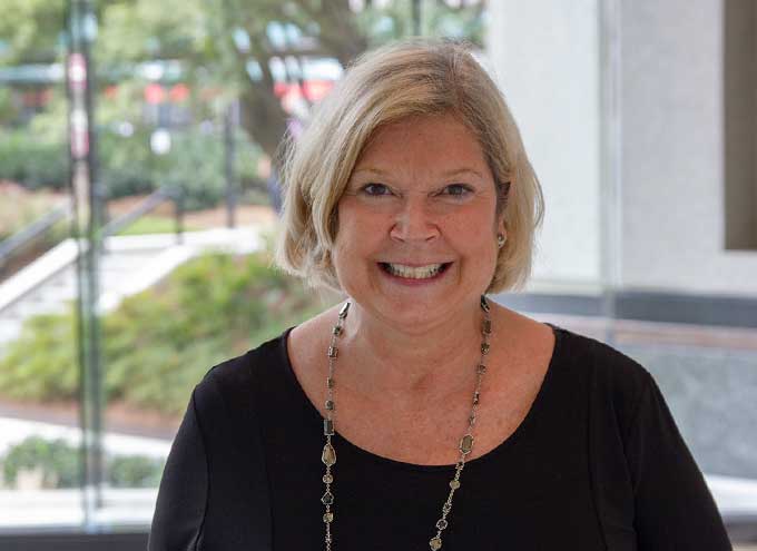 A woman with short blonde hair wearing a black top and a necklace stands indoors, smiling at the camera with greenery visible through a window behind her.