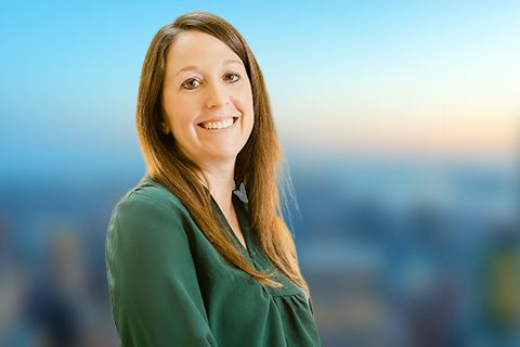 A woman with long brown hair wearing a green blouse smiles at the camera with a blurred cityscape background.