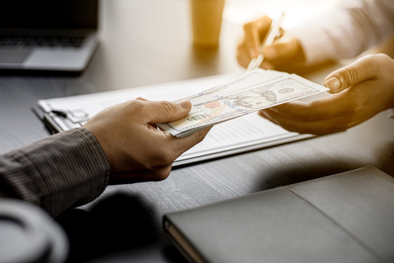 One person hands a stack of U.S. hundred-dollar bills to another person, who is signing a document on a desk with a laptop and notebook nearby.