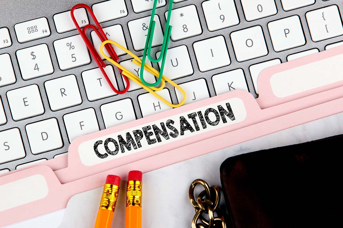 Folder labeled Compensation on a keyboard, with colorful paperclips, pencils, and part of a wallet and keychain visible.