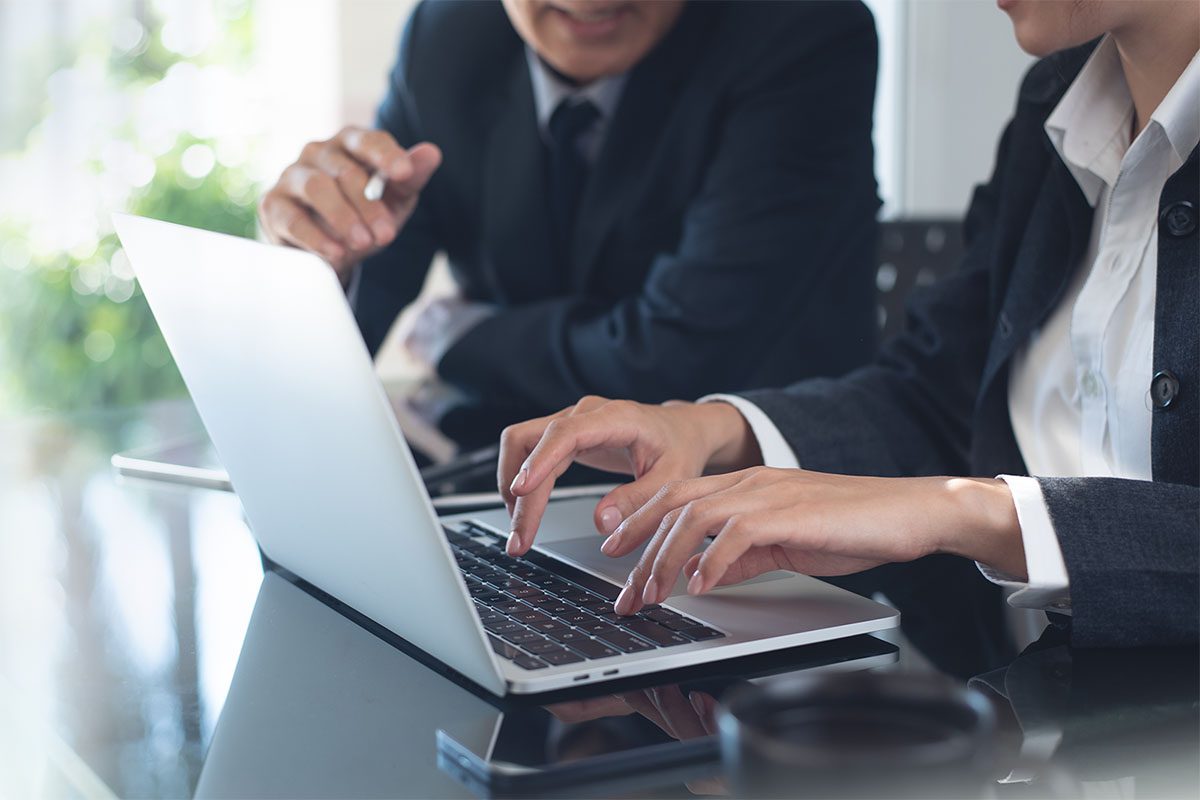 Two people in business attire sit at a table, one typing on a laptop while the other gestures with a pen, suggesting a work-related discussion.