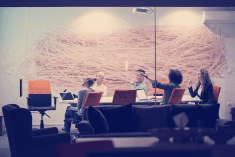 Five people sit around a conference table in a modern office meeting room, engaged in discussion. Laptops and papers are on the table, with abstract wall art in the background.