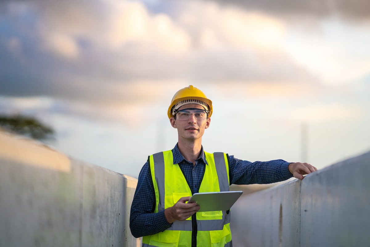 A man wearing a yellow safety vest, hard hat, and safety glasses stands outdoors holding a tablet at a construction site.