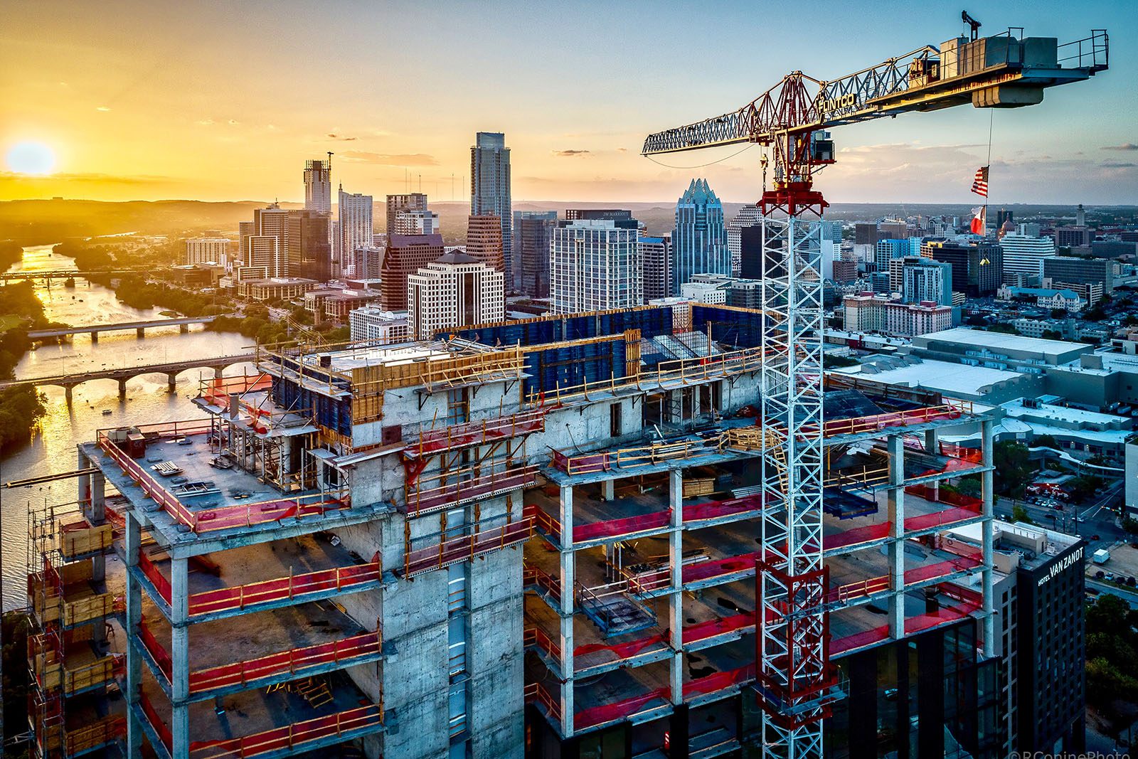 A high-rise building under construction with a large crane, set against a city skyline at sunset, with a river visible in the background.