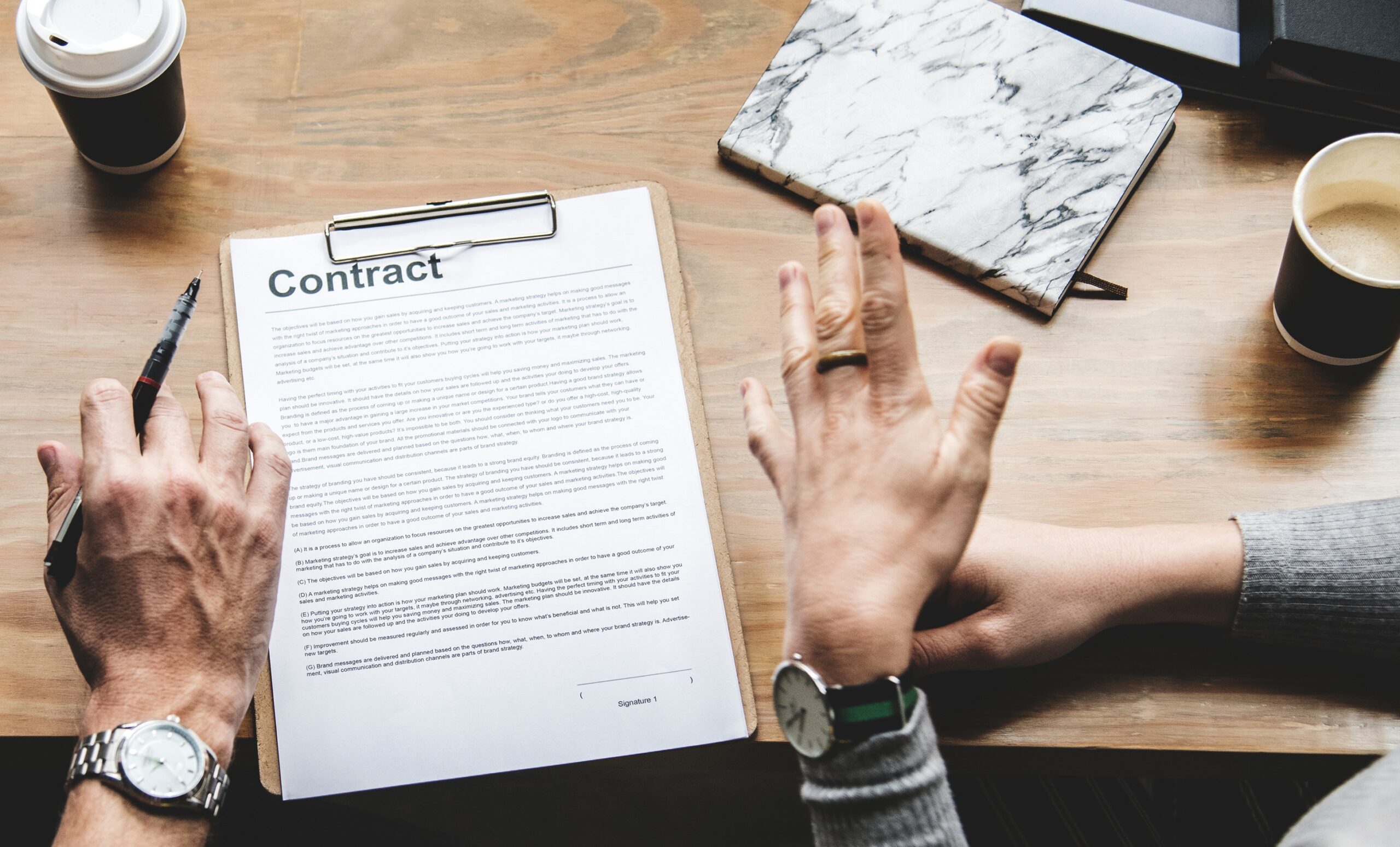 Two people are seated at a wooden table with coffee cups, discussing a contract document on a clipboard.