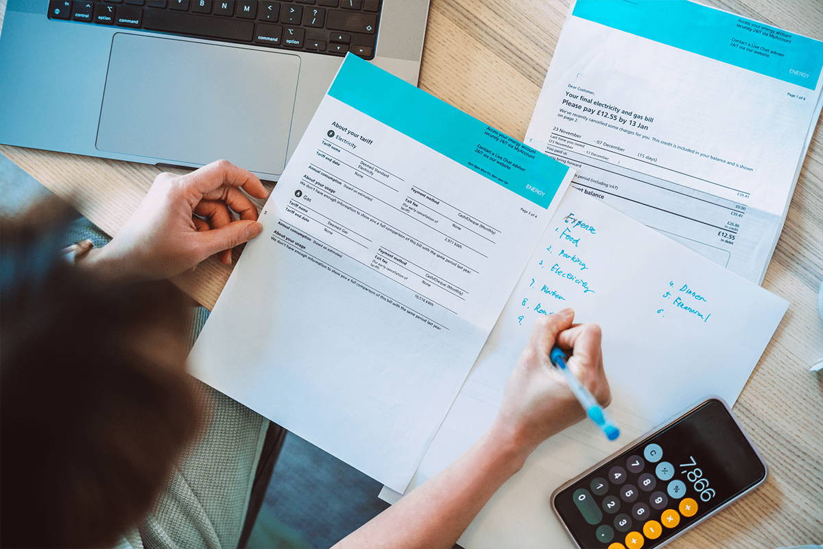 Person reviews documents and writes notes on paper next to a laptop, calculator, and several sheets with printed information on a desk.