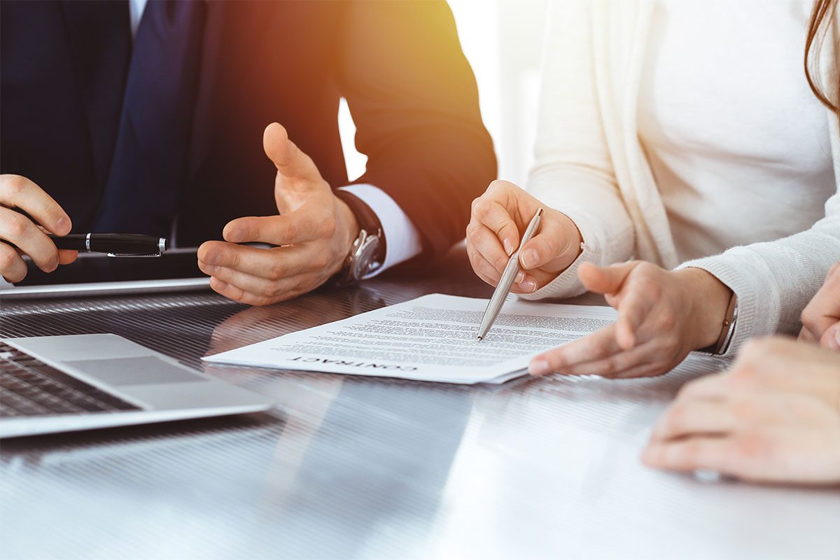 Three people sit at a table with a laptop, discussing and reviewing a contract document; one person points at the paper with a pen.
