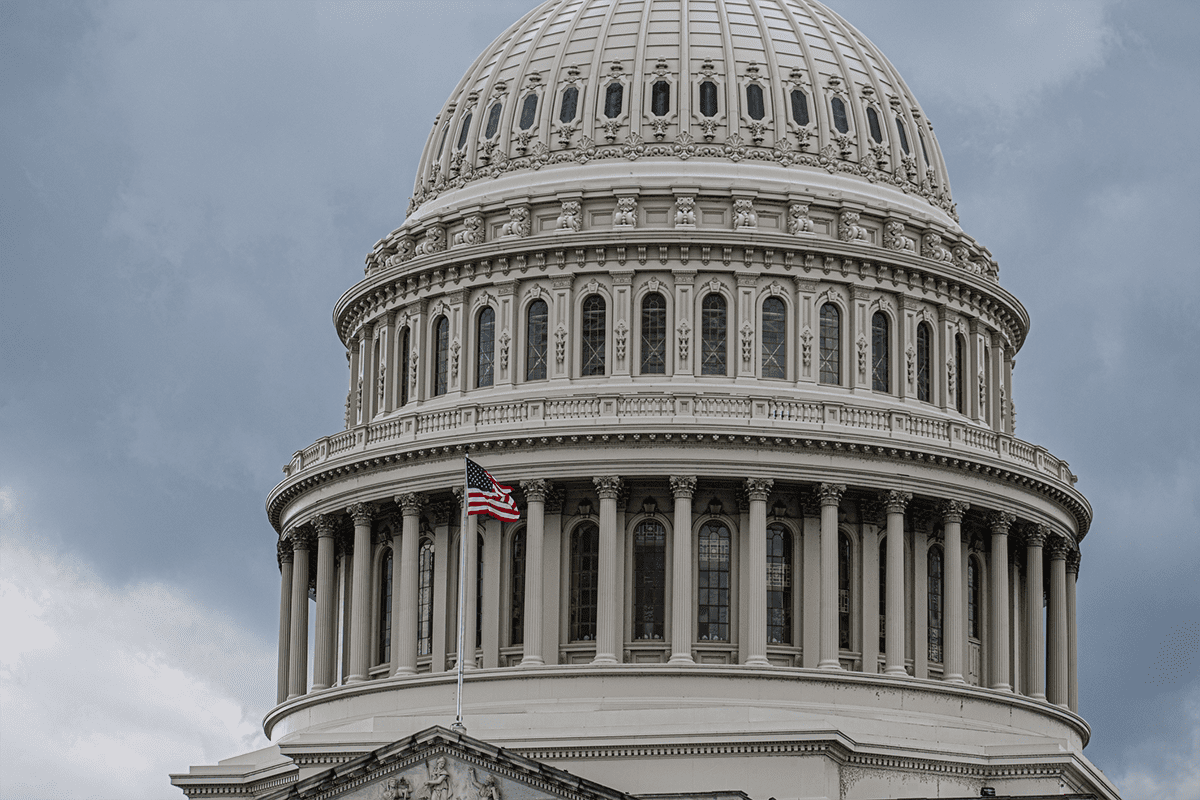 The dome of the United States Capitol building with an American flag flying in front, under a cloudy sky.