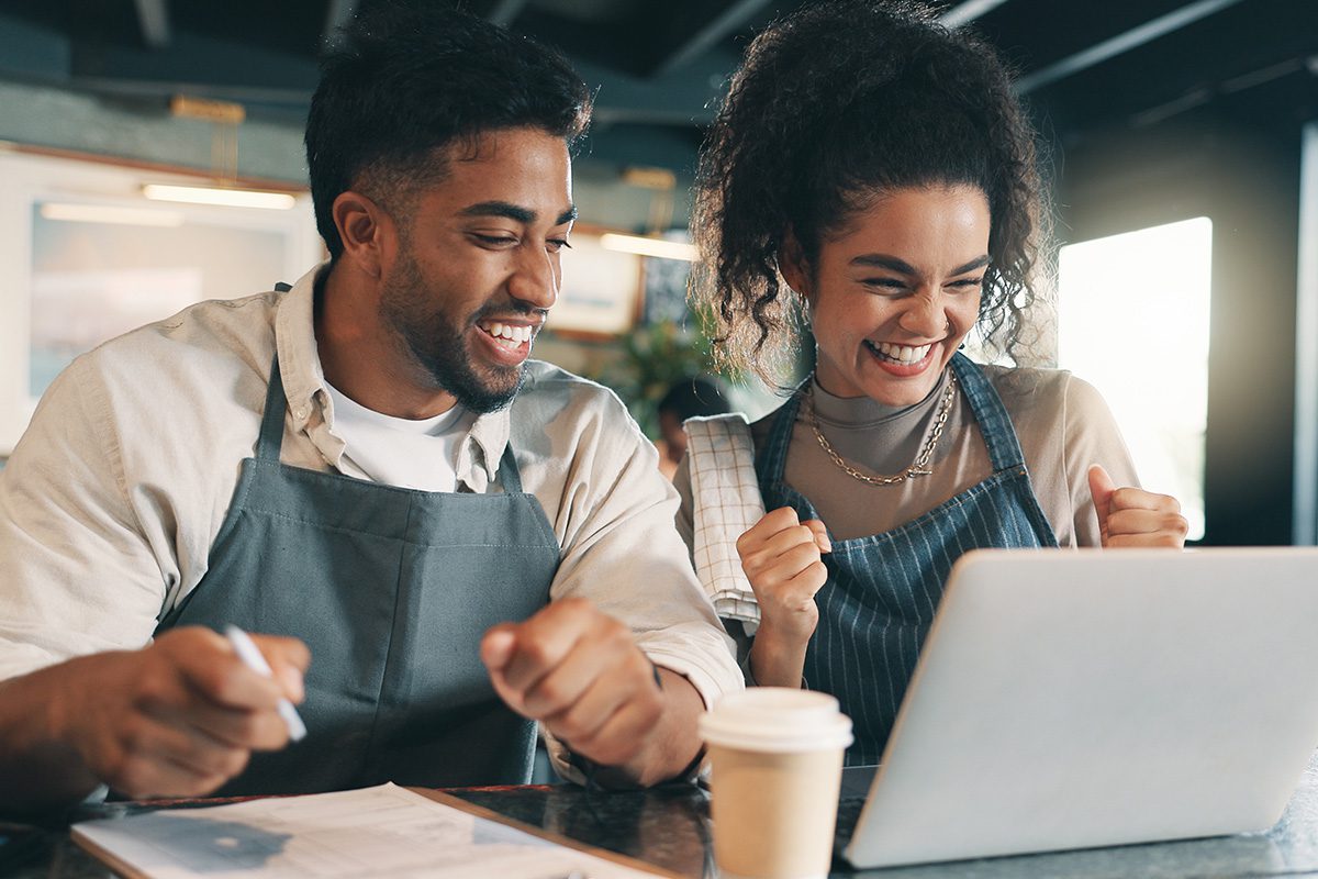 Two people wearing aprons sit at a table, smiling and cheering while looking at a laptop, with a coffee cup and notepad in front of them.