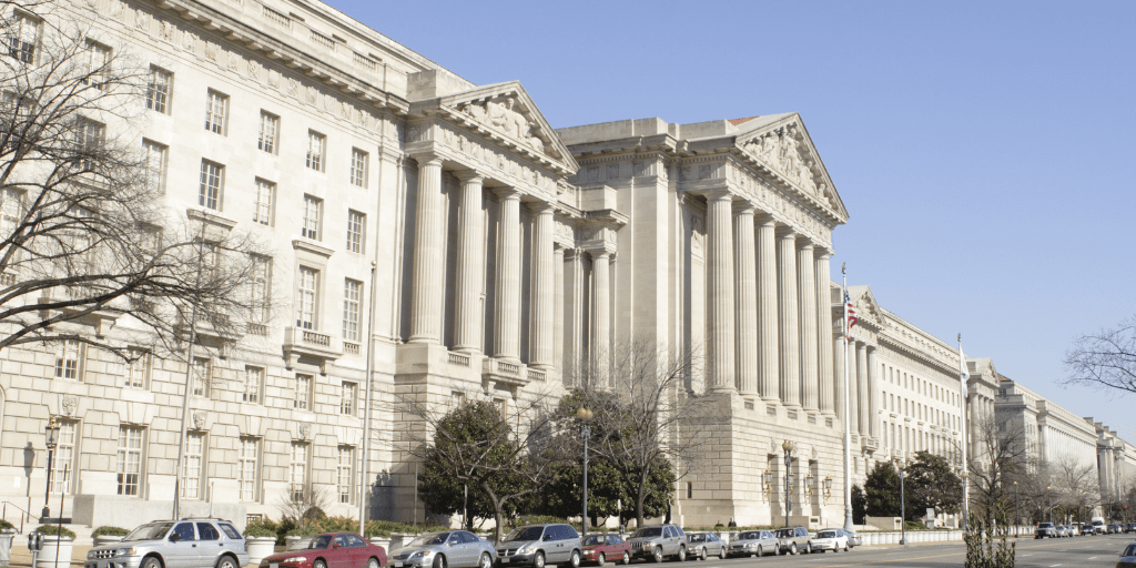 Large neoclassical government building with tall columns, multiple windows, and a row of parked cars along the street under a clear sky.