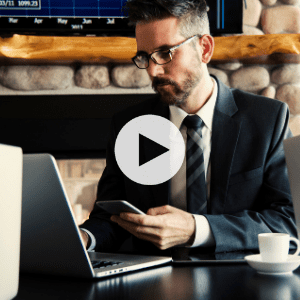 A man in a suit works at a desk with a laptop and smartphone; a cup and charts on a screen are visible in the background.