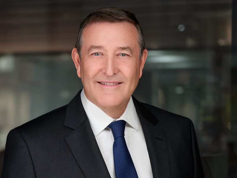 A middle-aged man in a dark suit, white shirt, and blue tie is smiling at the camera against a blurred office background.