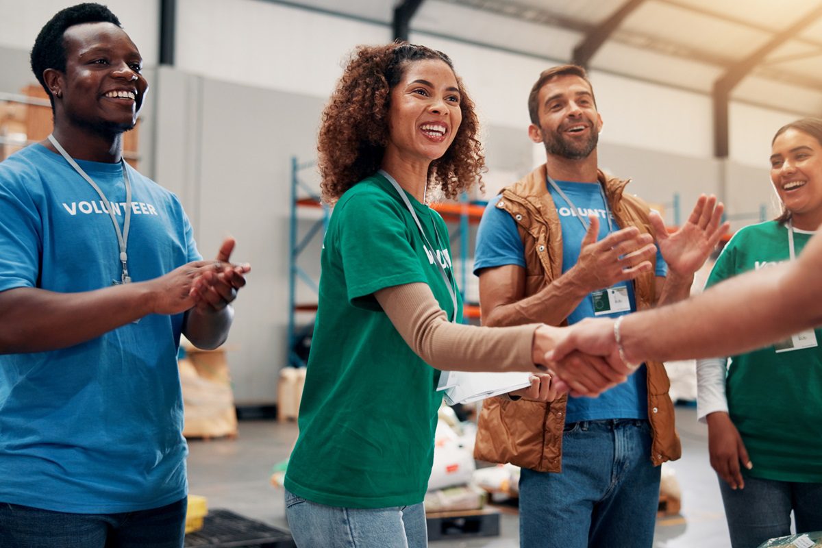 Four volunteers, wearing name tags and casual clothing, stand indoors smiling and clapping while one woman shakes hands with someone out of frame.