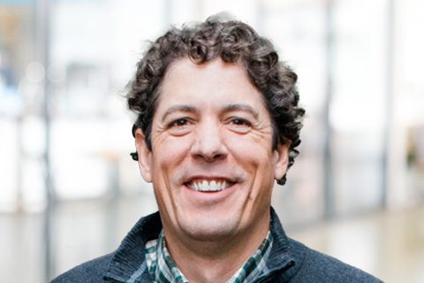 A man with short curly hair, wearing a plaid shirt and dark sweater, smiles at the camera in a brightly lit indoor setting.