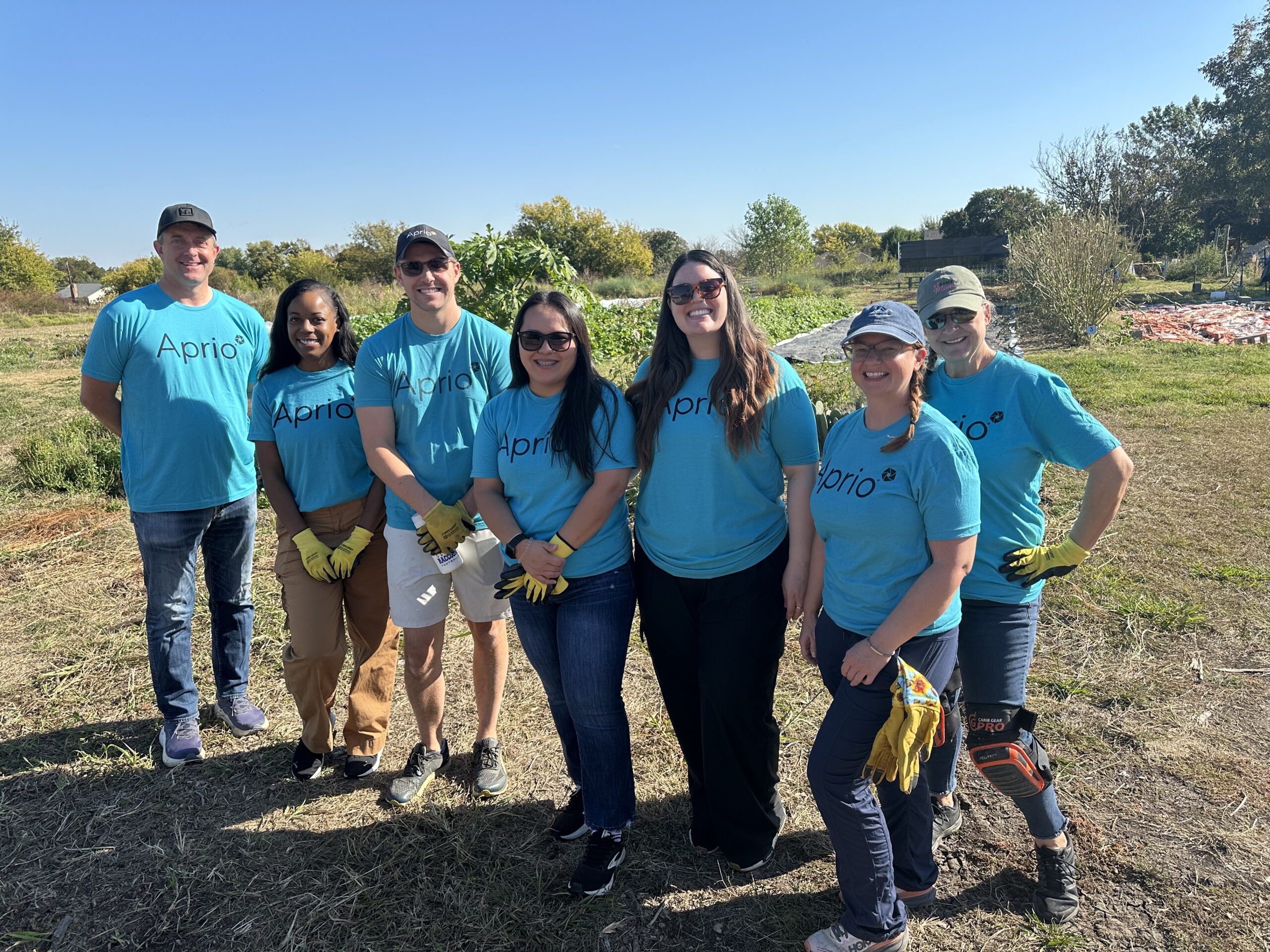 Seven people wearing matching blue Aprio shirts and yellow gloves stand together outdoors in a field on a sunny day.