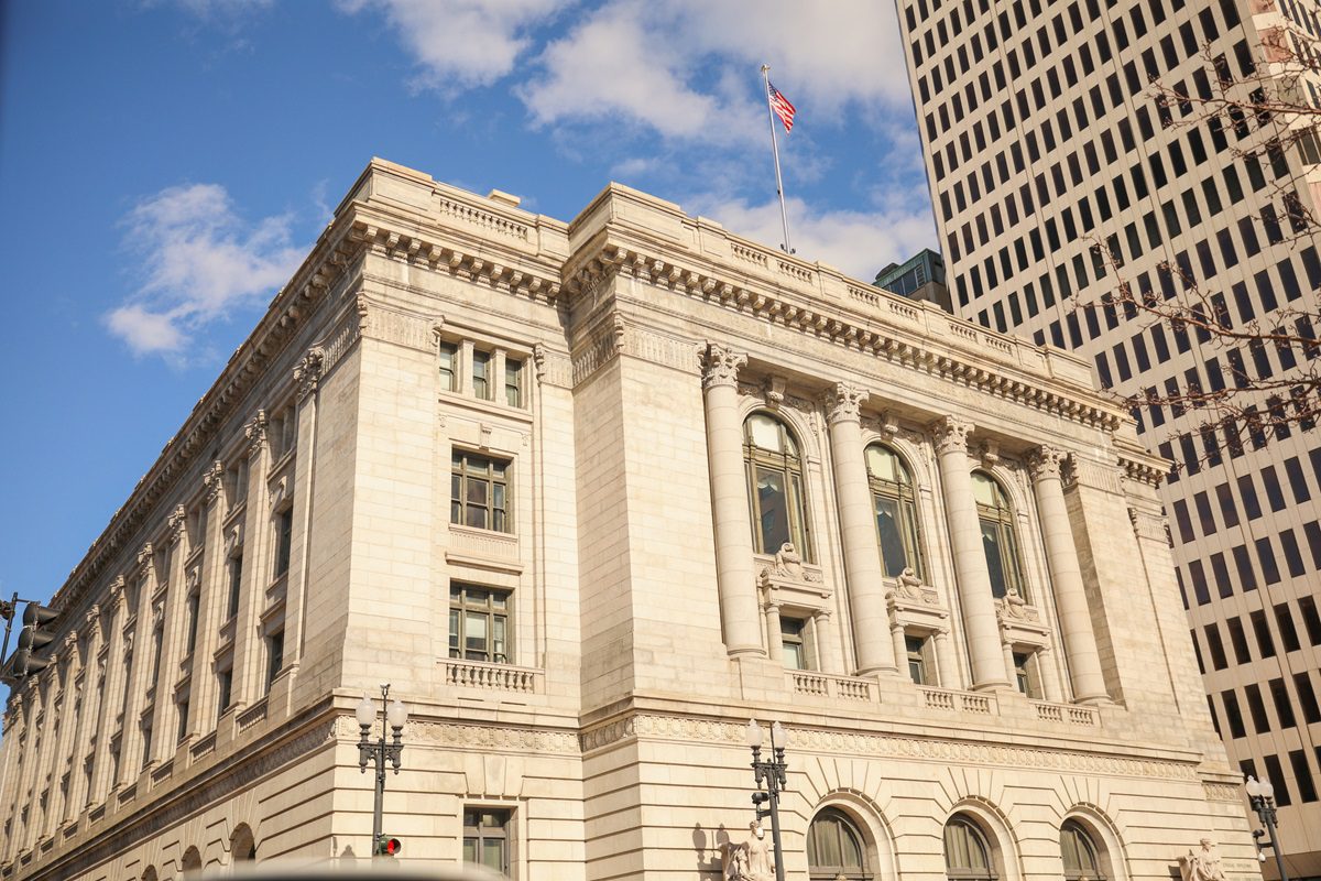 A historic stone building with columns and arched windows stands next to a modern high-rise, with an American flag flying on the roof under a blue sky.