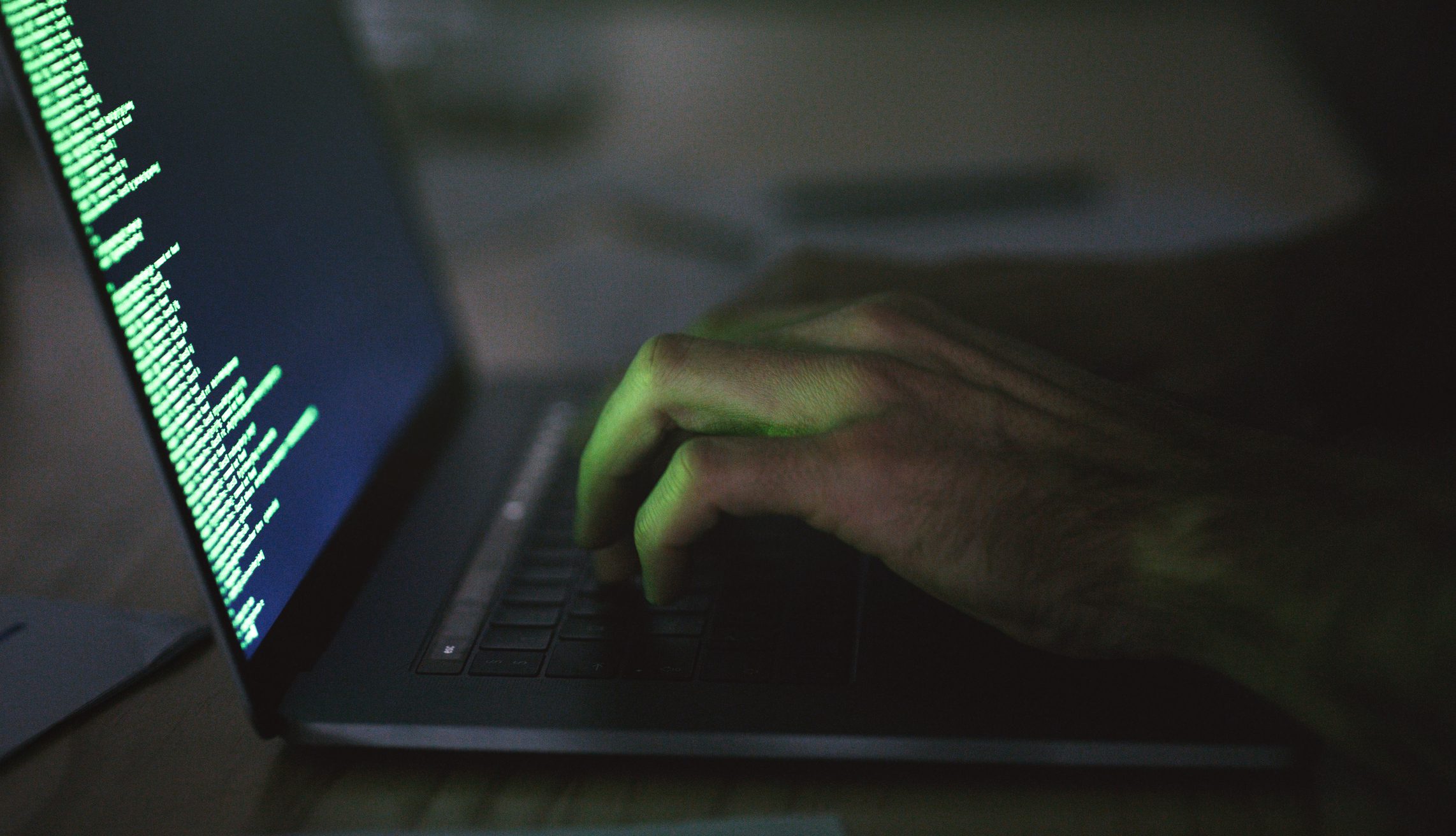 A person types on a laptop keyboard in a dimly lit room with green code displayed on the screen.