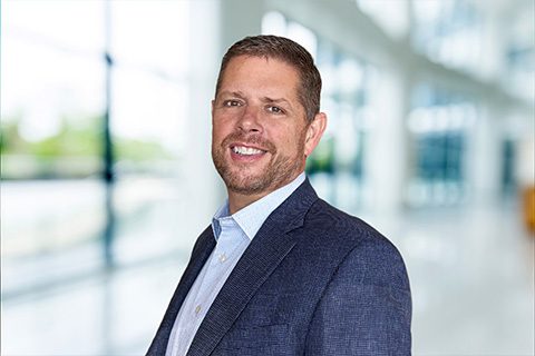 Dark haired man with a beard and dark blazer smiles in front of a bright blurred lobby background