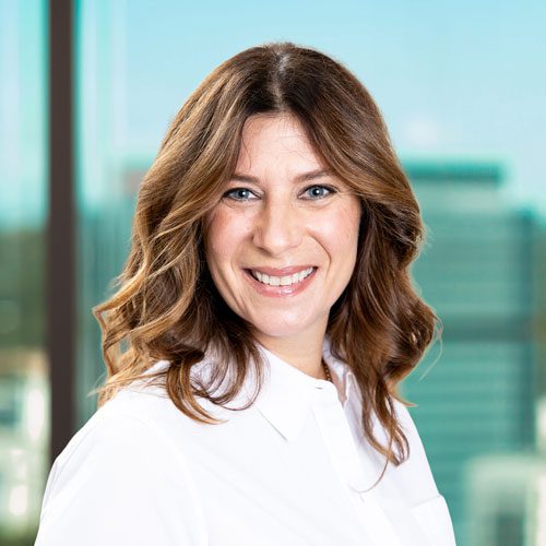 A woman with wavy brown hair and a white shirt smiles in front of a window with an urban cityscape in the background.