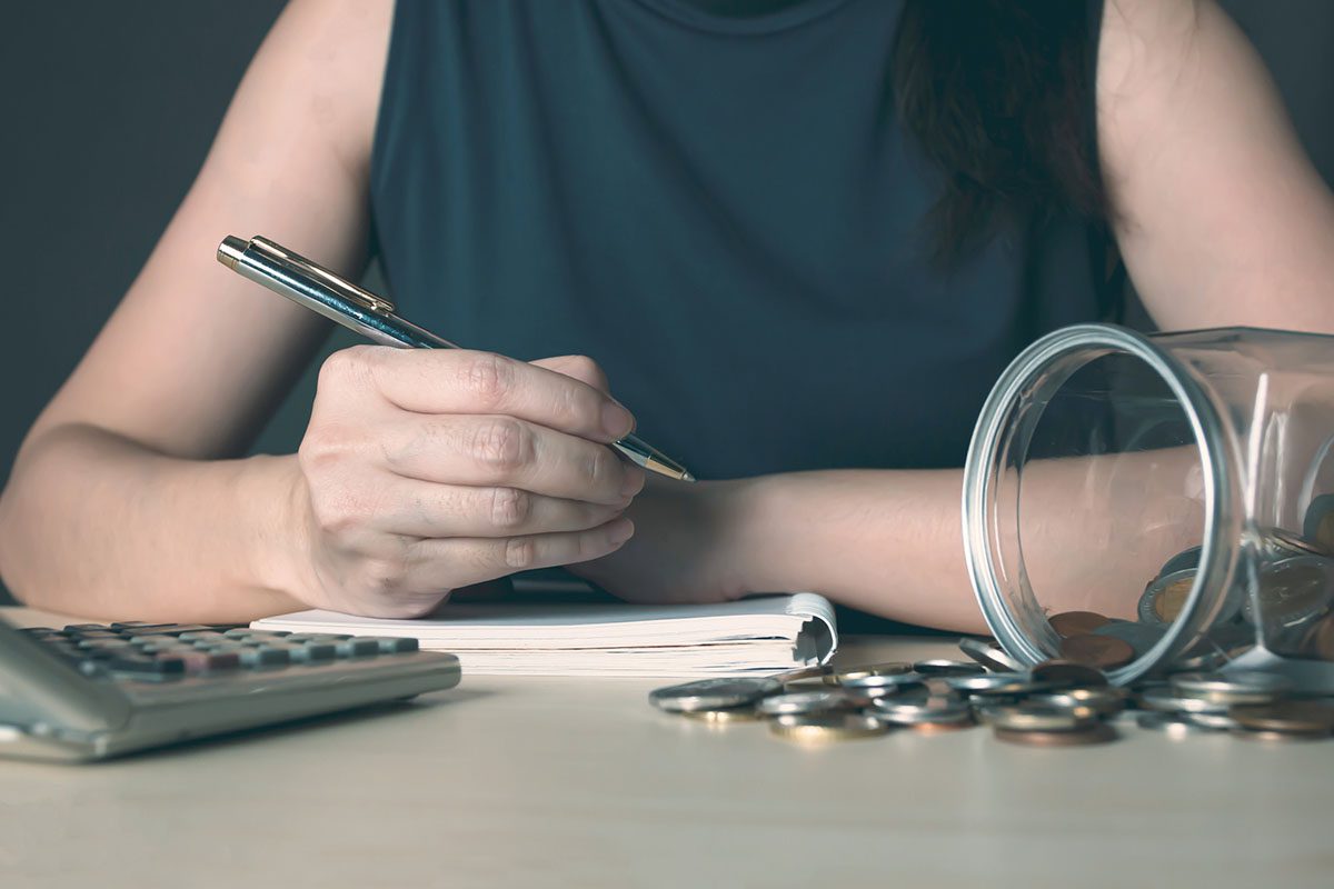 A person writes in a notebook beside a pile of coins, a tipped-over jar, and a calculator on a desk.