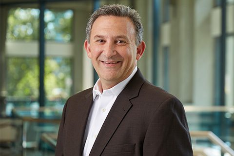A man in a dark suit and white shirt smiles while standing indoors in front of large windows with trees visible outside.