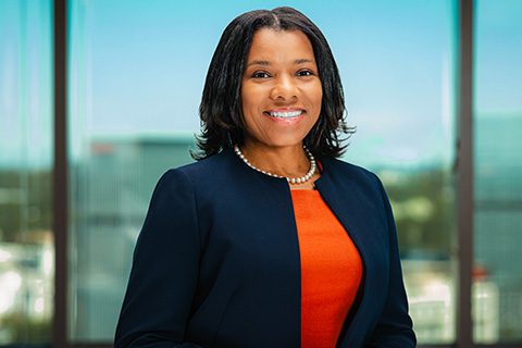 Dark haired woman in a navy jacket and red dress smiles in front of a bright window with a blurred city view.