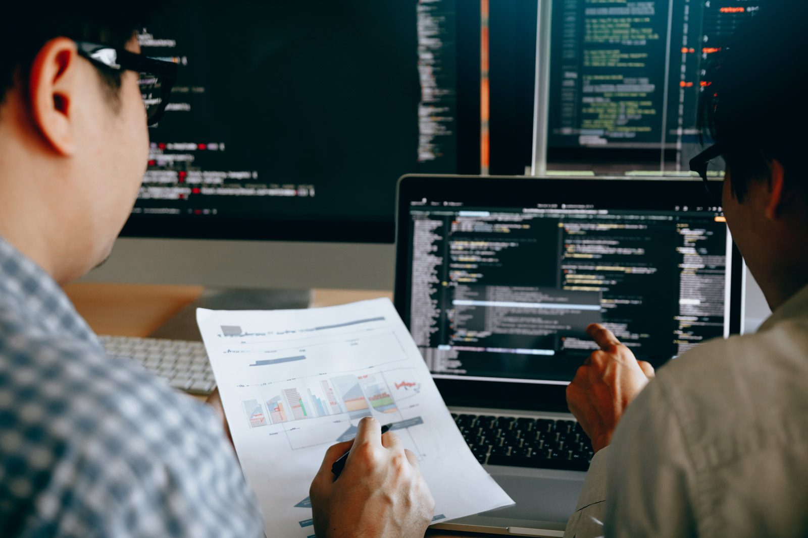 Two people review graphs on a printed report while analyzing source code on a laptop, with multiple monitors displaying code in the background.