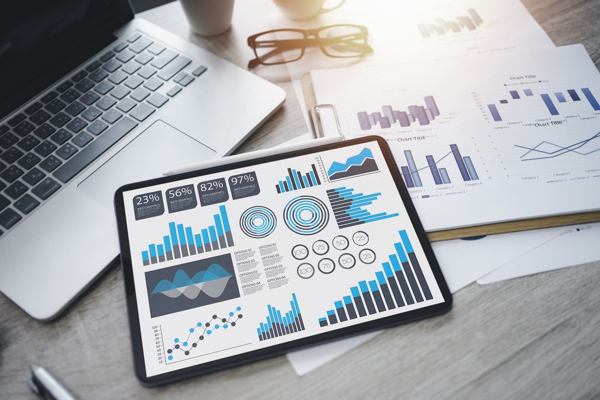 A tablet displays various blue and black charts and graphs on a desk next to a laptop, documents with similar graphs, a pen, and glasses.